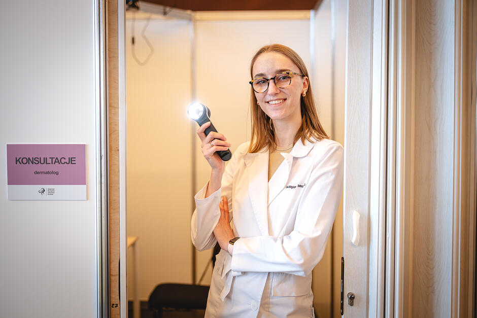 Lekarka w białym fartuchu stoi przed gabinetem, w ręku trzyma urządzenie medyczne do badania zmian skórnych. Na ścianie wisi tabliczka z napisem KONSULTACJE dermatolog  wraz z logo Gdańskiego Kongresu Zdrowia Kobiet
