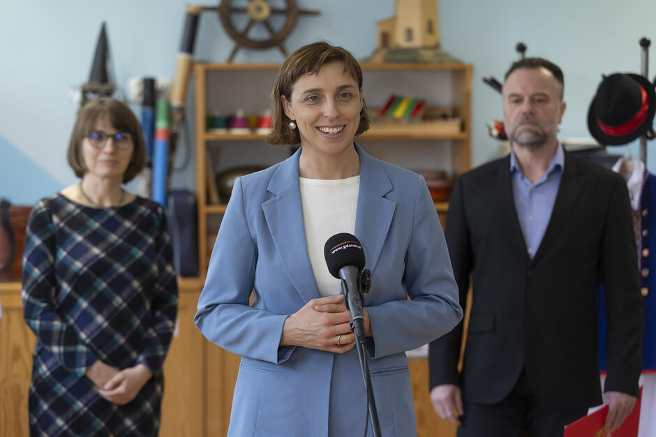 Na zdjęciu widać uśmiechniętą kobietę w błękitnym kostiumie, która przemawia do mikrofonu oznaczonego logo Gdańska. W tle, w pomieszczeniu przypominającym salę przedszkolną lub szkolną, stoją dwie inne osoby, a na regale widoczne są morskie dekoracje, takie jak koło sterowe i model latarni morskiej. 