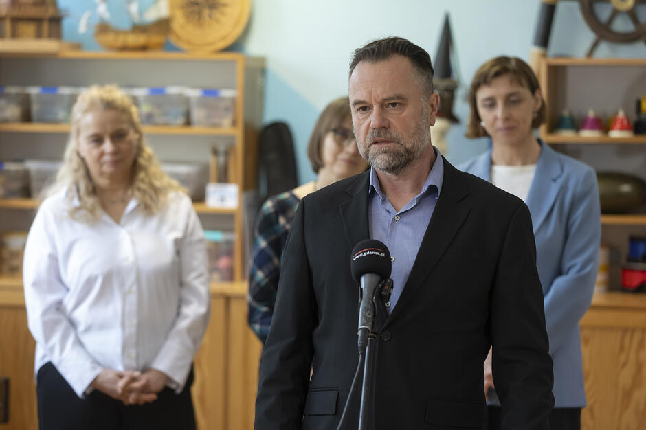 Na zdjęciu widać brodatego mężczyznę w ciemnej marynarce, który przemawia do mikrofonu z logo Gdańska podczas oficjalnego wydarzenia. W tle, w pomieszczeniu o charakterze edukacyjnym, stoją dwie kobiety, a na regałach widoczne są morskie dekoracje, takie jak modele statków i kół sterowych.
