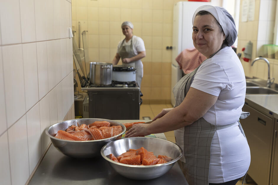 Zdjęcie przedstawia uśmiechniętą kobietę w białym czepku i fartuchu, która przygotowuje surowe filety łososia w profesjonalnej kuchni. W tle widać drugą pracownicę oraz wyposażenie kuchenne, takie jak duże garnki, zlewozmywak i wykafelkowane ściany.