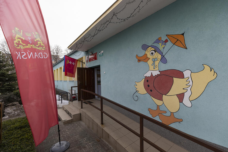 Zdjęcie przedstawia wejście do Przedszkole nr 60 im. Jana Brzechwy w Gdańsku, które wyróżnia się niebieską elewacją ozdobioną kolorowym muralem przedstawiającym uśmiechniętą kaczkę. Przed budynkiem widoczna jest czerwona flaga z herbem Gdańska oraz podjazd dla wózków prowadzący do drzwi wejściowych.