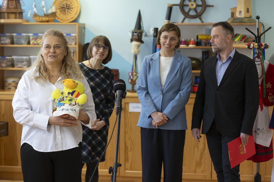 Na pierwszym planie uśmiechnięta kobieta w białej koszuli prezentuje pluszową żółtą kaczkę ozdobioną licznymi przypinkami, co może nawiązywać do słynnej „Kaczki Dziwaczki”.