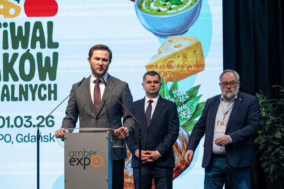 Mężczyzna przemawia do mikrofonu, za nim dwóch innych mężczyzn na scenie, za nimi ekran.