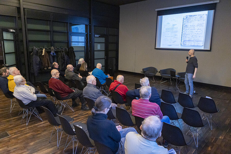 Sala z rzędami krzeseł na których siedzi kilkanaście osób. Na ścianie w głębi duży ekran pod którym stoi przemawiający mężczyzna z mikrofonem