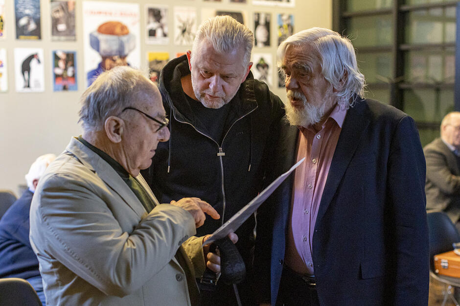 Trzej mężczyźni stoją rozmawiając - pośrodku mężczyzna w średnim wieku - po lewej i prawej mężczyźni w wieku senioralnym 