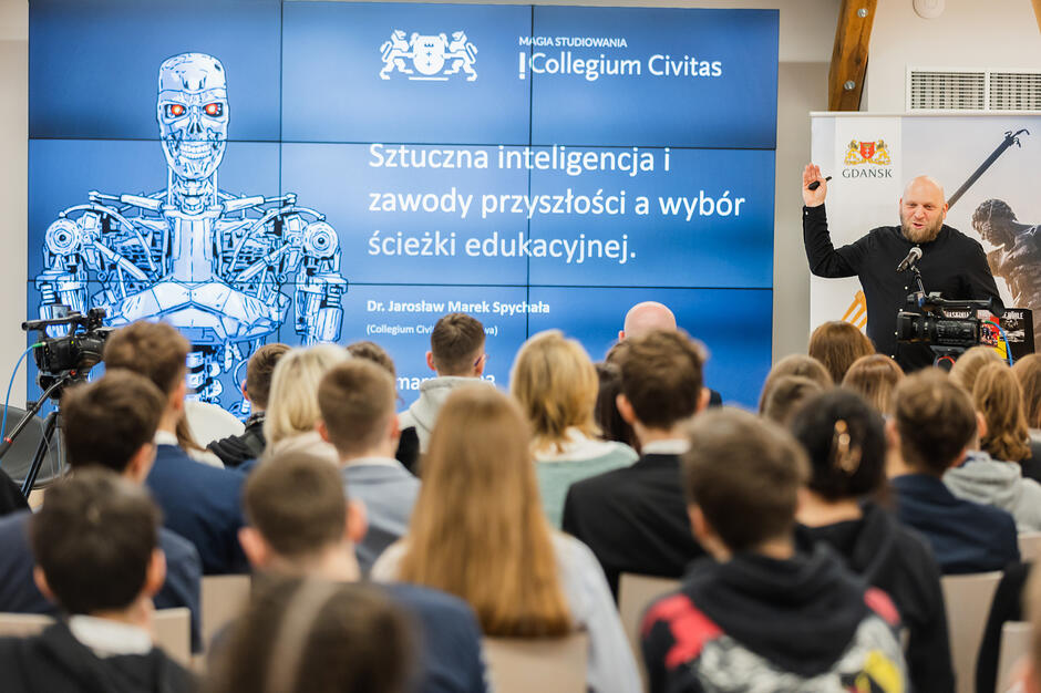 Sala wypełniona młodzieżą do której przemawia łysy mężczyzna z brodą