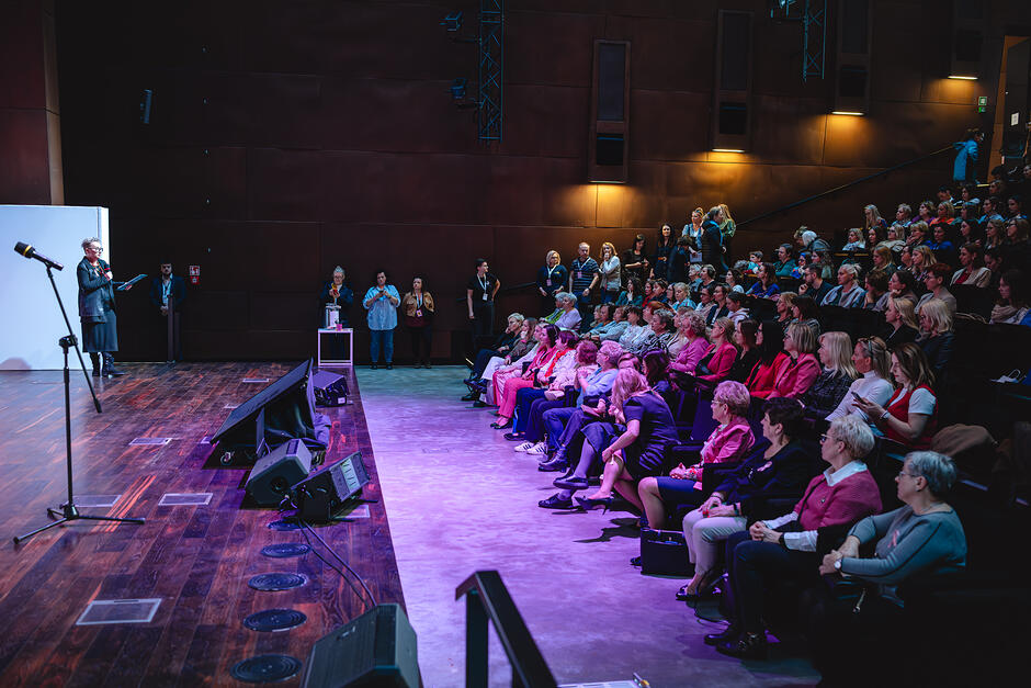Na zdjęciu widać salę audytoryjną Europejskiego Centrum Solidarności. Po lewej stronie, na drewnianej scenie, stoi osoba przemawiająca do mikrofonu, trzymająca kartki lub notatki. Obok znajduje się statyw z mikrofonem oraz sprzęt nagłośnieniowy ustawiony na podłodze.
Po prawej stronie siedzi publiczność w rzędach podwyższonych miejsc. Większość uczestników to kobiety w różnym wieku, ubrane w stroje codzienne lub półformalne. Część osób patrzy w stronę sceny, inne rozmawiają lub obserwują otoczenie. W tle stoją także osoby przy ścianie.
Wnętrze ma ciemne, brązowe ściany i nastrojowe oświetlenie – scena jest oświetlona jaśniej, a widownia częściowo pogrążona w półmroku z fioletowo-niebieskimi akcentami świetlnymi.