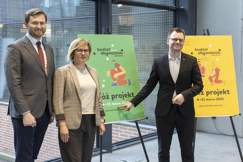 na zdjęciu dwóch mężczyzn i jedna kobieta, wszyscy w oficjalnych garniturach, pozują do zdjęcia przy kolorowych tablicach