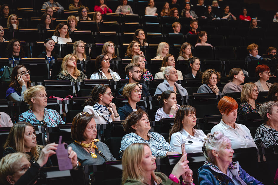 Na zdjęciu widać salę audytoryjną Europejskiego Centrum Solidarności. W rzędach podwyższonych miejsc siedzi publiczność- większość to kobiety w różnym wieku, ubrane w stroje codzienne lub półformalne