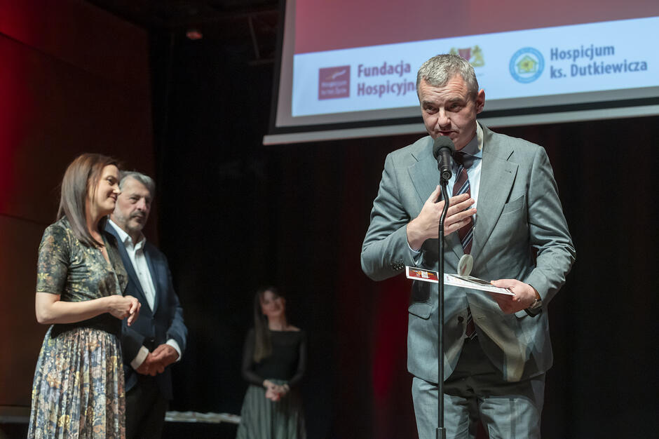 Na zdjęciu widać mężczyznę w garniturze przemawiającego do mikrofonu, który trzyma w dłoniach dokumenty lub nagrodę. W tle znajduje się ekran z logotypami Fundacji Hospicyjnej oraz Hospicjum ks. Dutkiewicza w Gdańsku