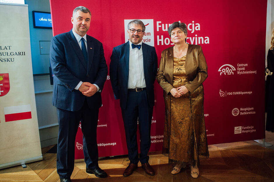 Na zdjęciu widać trzy elegancko ubrane osoby pozujące na tle czerwonej ścianki z logotypami Fundacji Hospicyjnej oraz Hospicjum ks. Dutkiewicza. Pośrodku stoi mężczyzna w okularach i granatowej muszce, a obok niego kobieta w długiej, złotej sukni wieczorowej z narzutką.