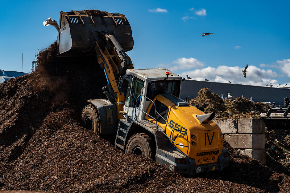 Na pierwszym planie ciężki sprzęt w kolorze czarno-żółtym, który pracuje z górą ciemnej ziemi