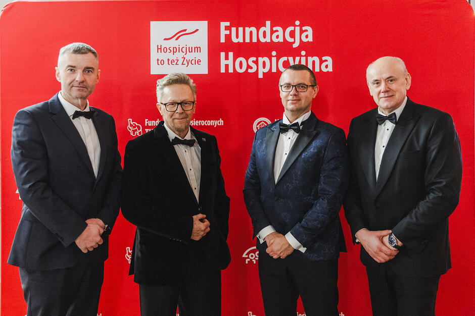 Na zdjęciu widać czterech mężczyzn w eleganckich garniturach i ciemnych muszkach, którzy pozują razem do wspólnej fotografii. Stoją oni na tle intensywnie czerwonej ścianki z logotypami Fundacji Hospicyjnej oraz hasłem „Hospicjum to też Życie”.
