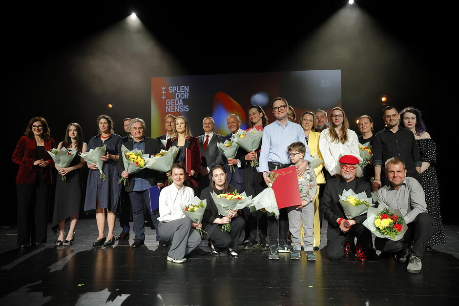 Grupa laureatów i organizatorów stoi wspólnie na scenie podczas uroczystej gali wręczenia nagród Splendor Gedanensis. Uczestnicy trzymają bukiety kwiatów oraz teczki z wyróżnieniami, a za nimi widoczny jest duży ekran z nazwą wydarzenia. Całość tworzy elegancką, podniosłą atmosferę, podkreślając charakter ceremonii i celebrację osiągnięć nagrodzonych osób