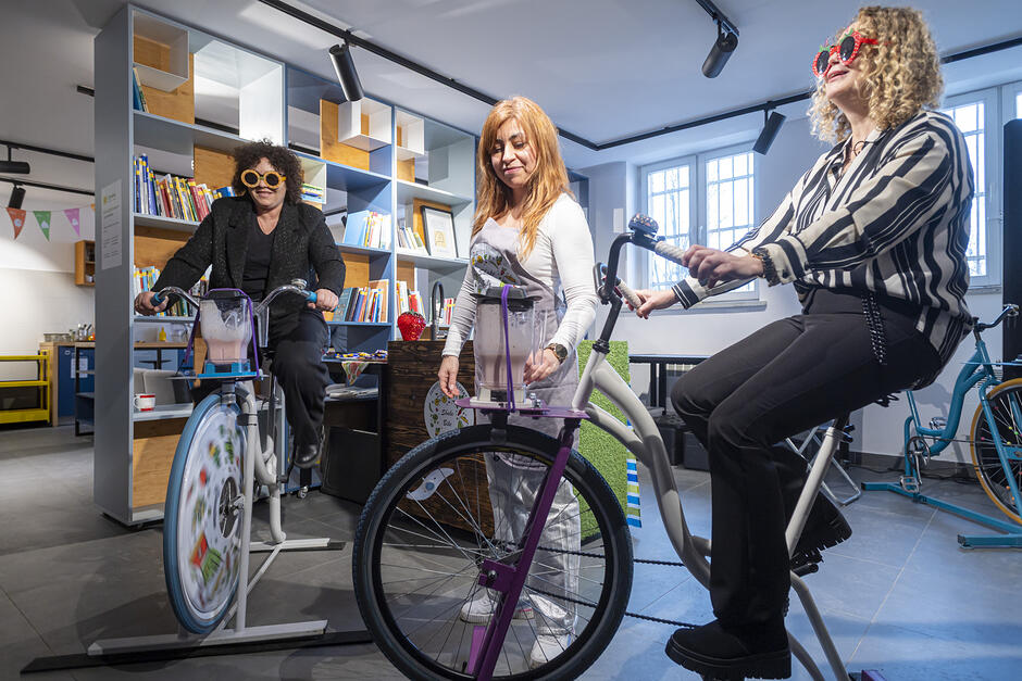 na zdjęciu dwie kobiety na rowerach stacjonarnych, mają zabawne okulary przeciwsłoneczne, między nimi stoi trenerka