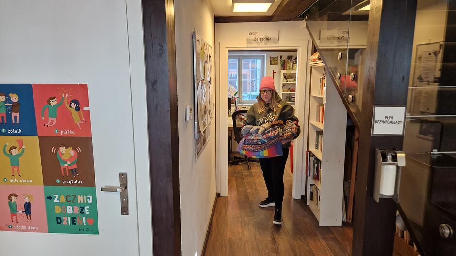 Wnętrze domu. Po lewej białe drzwi, po prawej schody, pośrodku dziewczyna w różowej wełnianej czapce i okularach z naręczem czapek i szalików