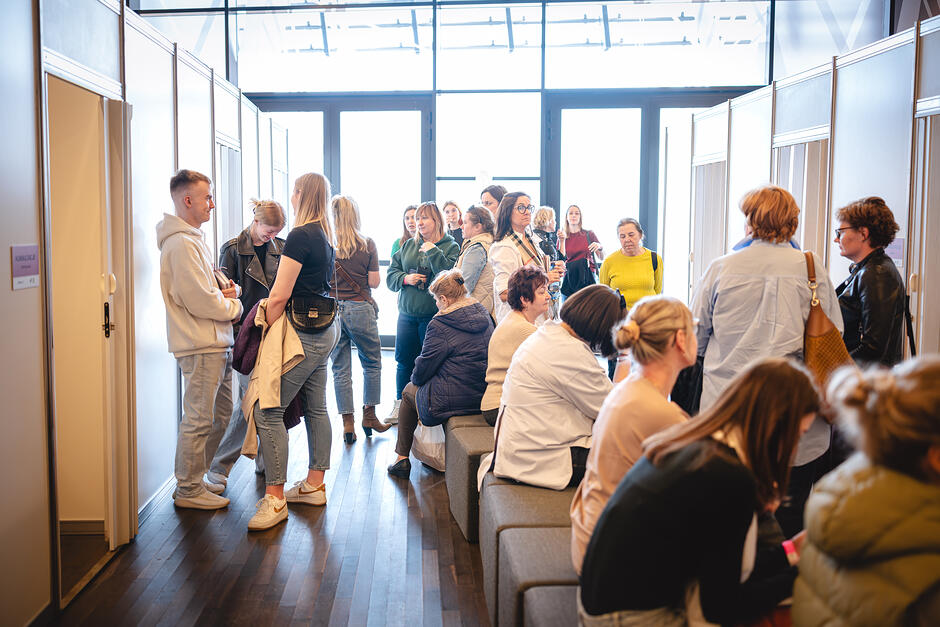 Na zdjęciu widać grupę osób stojących i siedzących w jasnym korytarzu z przeszklonym wejściem na końcu. Po obu stronach znajdują się boksy, w których odbywały się konsultacje i badania lekarskie podczas Gdańskiego Kongresu Zdrowia Kobiet w 2025. Część osób rozmawia ze sobą, inne siedzą na ławkach, trzymając torebki i okrycia wierzchnie.