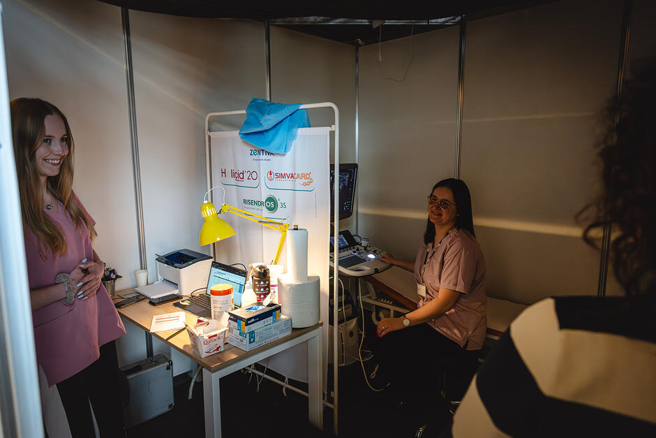 Na zdjęciu widać dwie uśmiechnięte kobiety- obie w lekarskich fartuchach. Kobiety patrzą na pacjentkę, która wchodzi do zaciemnionego pomieszczenia- gabinetu USG. Jedna z kobiet siedzi przy urządzeniu diagnostycznym i leżance, odgrodzonych parawanem, druga stoi przy stoliku, na którym znajdują się komputer, drukarka oraz przybory medyczne.