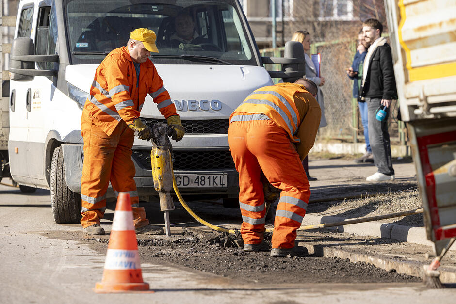 Dwóch robotników budowlanych w pomarańczowych kombinezonach naprawiają nawierzchnię ulicy, jeden trzyma dużą wiertarkę