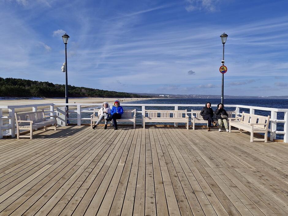 Na pierwszym planie znajdują się białe ławki stojące na drewnianym podeście. Są ustawione obok siebie i naprzeciwko siebie, jest ich łącznie pięć. Na dwóch z nich siedzą ludzie. W tle widać plażę i wodę