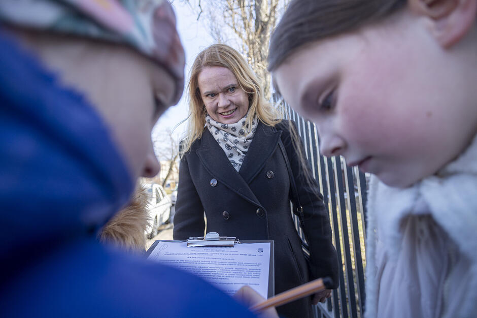 Na zdjęciu widać uśmiechniętą kobietę w ciemnym płaszczu, która obserwuje dwoje dzieci pochylonych nad dokumentem na podkładce z klipsem. Jedno z dzieci trzyma długopis, co sugeruje, że wspólnie rozwiązują zadanie lub wypełniają ankietę na świeżym powietrzu.