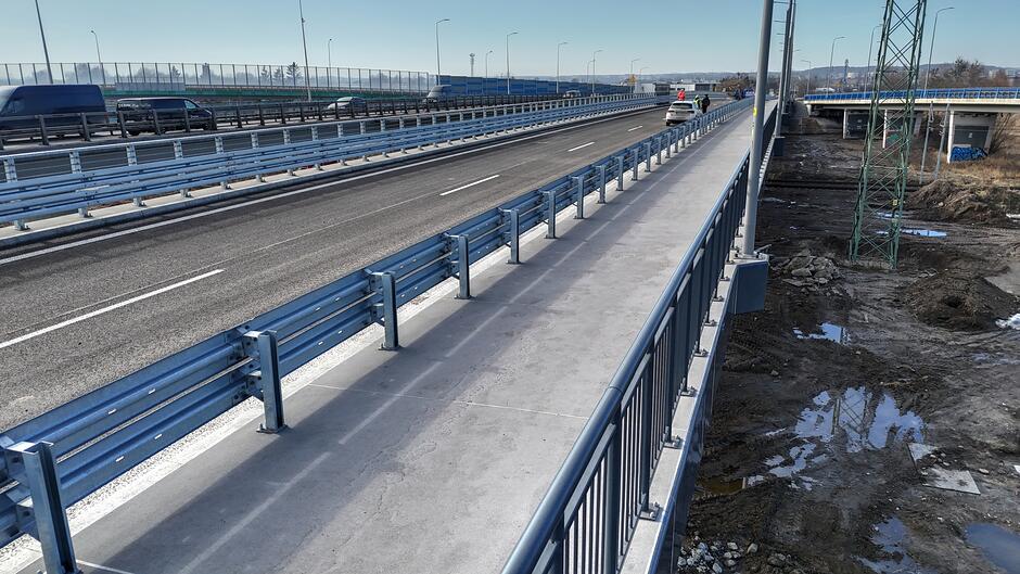 Na zdjęciu widać nowo wybudowany lub wyremontowany odcinek drogi z wiaduktem, wyposażony w lśniące, metalowe barierki energochłonne i chodnik dla pieszych. W oddali dostrzec można poruszające się samochody oraz infrastrukturę techniczną, taką jak słupy oświetleniowe i kratownicowy maszt.