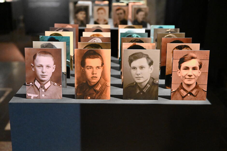 Zdjęcie przedstawia wystawę „Nasi chłopcy” w Muzeum Gdańska, prezentującą rzędy czarno-białych i sepia portretów młodych mężczyzn w mundurach z czasów II wojny światowej. Na pierwszym planie widoczne są sylwetki osób takich jak Zygmunt Ostrowski, których losy ilustrują tragiczne historie przymusowego wcielenia Polaków do Wehrmachtu.