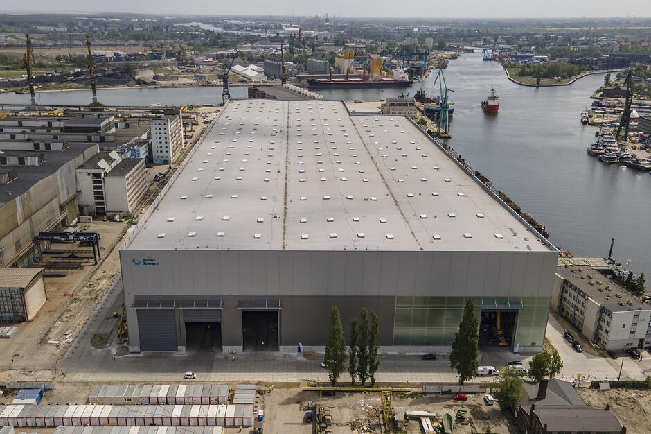 Zdjęcie przedstawia nowoczesną halę produkcyjną firmy Baltic Towers, położoną na terenie stoczniowym w Gdańsku bezpośrednio nad Martwą Wisłą. W tle widoczny jest industrialny krajobraz portowy z charakterystycznymi żurawiami, statkami oraz infrastrukturą przemysłową nabrzeża.

