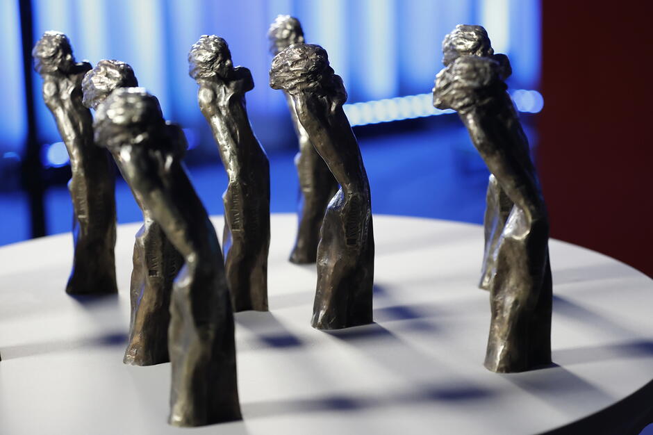 Na zdjęciu widać grupę mosiężnych statuetek ustawionych na białym, okrągłym blacie, które przedstawiają stylizowaną postać mężczyzny wyłaniającego się z fal
