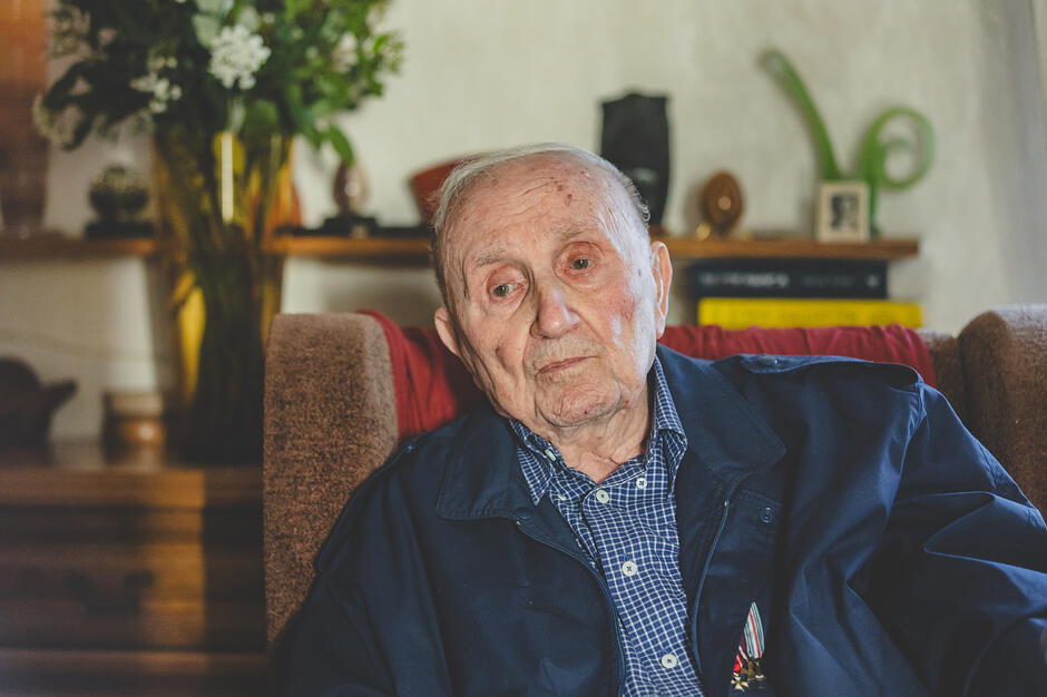  starszy mężczyzna siedzi na kanapie, ma na sobie niebieską kurtkę z czerwono-białą koszulą z kratkami pod spodem, mężczyźni mają poważny wyraz twarzy i patrzą bezpośrednio na kamerę, w tle znajdują się różne przedmioty dekoracyjne, takie jak wazon z białymi kwiatami, zegar i ramka obrazowa na ścianie.