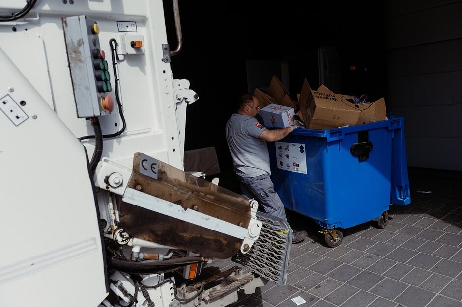 Na zdjęciu widać pracownika komunalnego opróżniającego duży, niebieski kontener wypełniony kartonowymi opakowaniami. Mężczyzna stoi obok białej śmieciarki, przygotowując odpady do załadunku w miejskiej scenerii.
