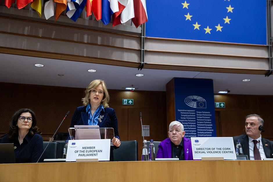 Kobieta stoi na mównicy w sali obrad i przemawia, obok siedzą dwie inne kobiety i mężczyzna.