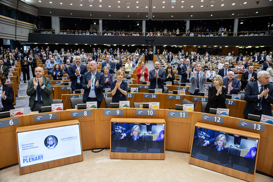 W dużej sali obrad ludzie stoją i bija brawo.