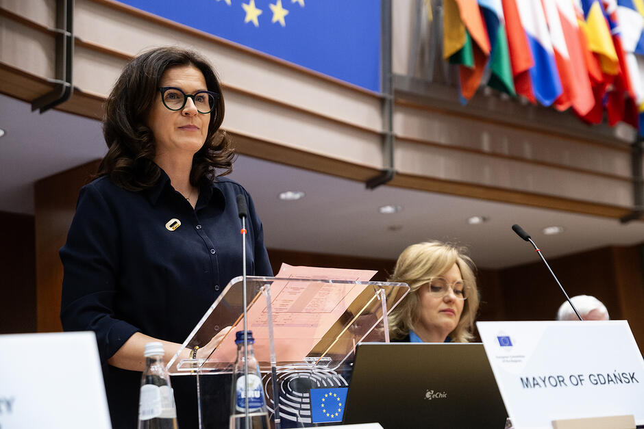 Kobieta stoi na mównicy w sali obrad i przemawia.