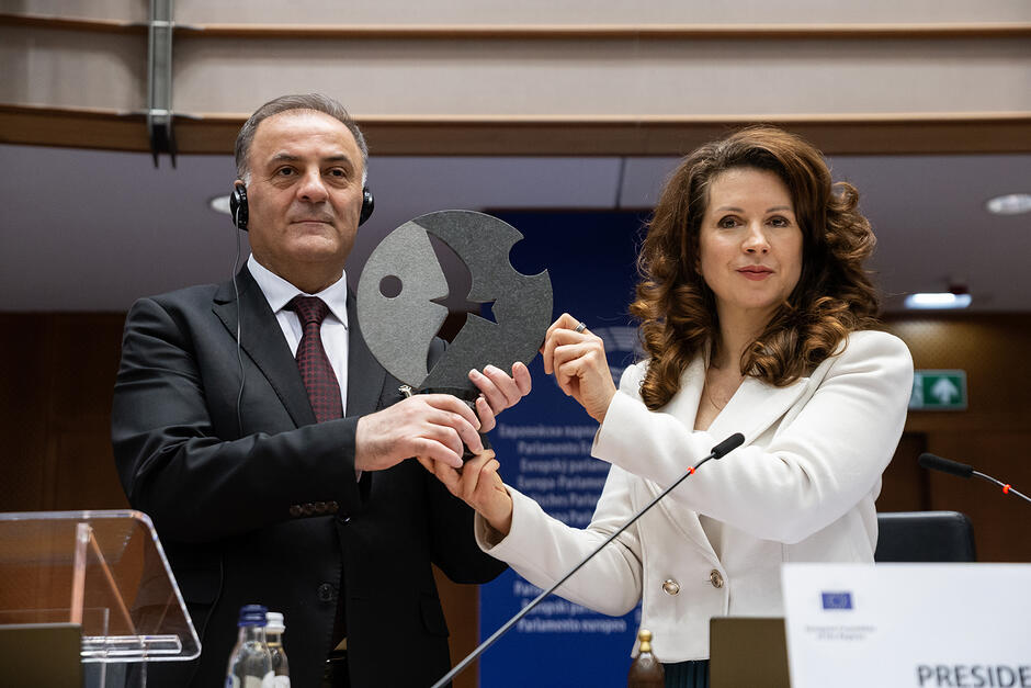 Kobieta i mężczyzna stoją na mównicy sali obrad i trzymają statuetkę.