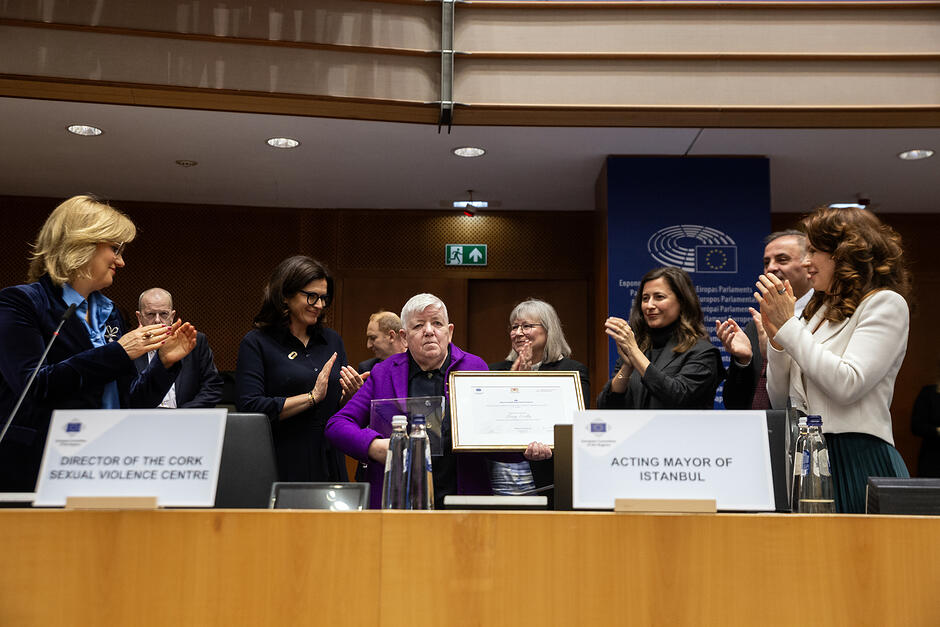 Kobieta na mównicy w sali obrad trzyma dyplom i statuetkę, otacza ją grono osób, które biją brawo.