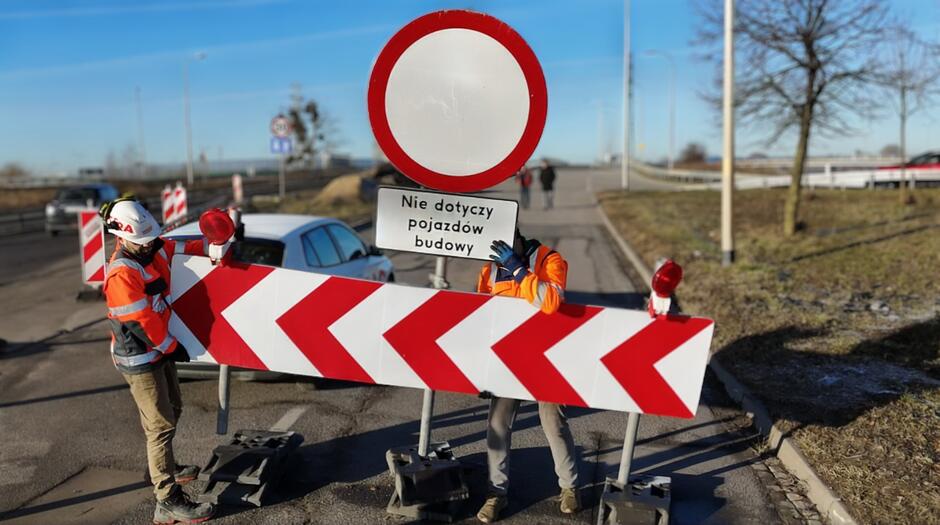 na zdjęciu białoczerwony znak drogowy, trzyma go dwóch robotników