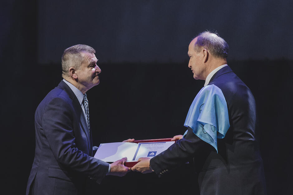 Na zdjęciu widać uroczystość wręczenia dyplomu lub nagrody, podczas której mężczyzna po lewej stronie przekazuje otwarte etui osobie w ciemnym garniturze. Mężczyzna odbierający wyróżnienie ma przewieszoną przez ramię błękitną chustę z logo, co sugeruje oficjalny charakter wydarzenia