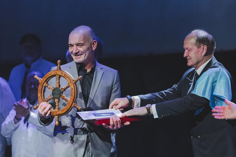 Zdjęcie przedstawia radosny moment wręczenia nagrody, na którym mężczyzna w szarym garniturze trzyma pamiątkowe drewniane koło sterowe. Obok niego stoi drugi mężczyzna z błękitną chustą na ramieniu, wspólnie prezentując dokument lub certyfikat w ozdobnym etui.