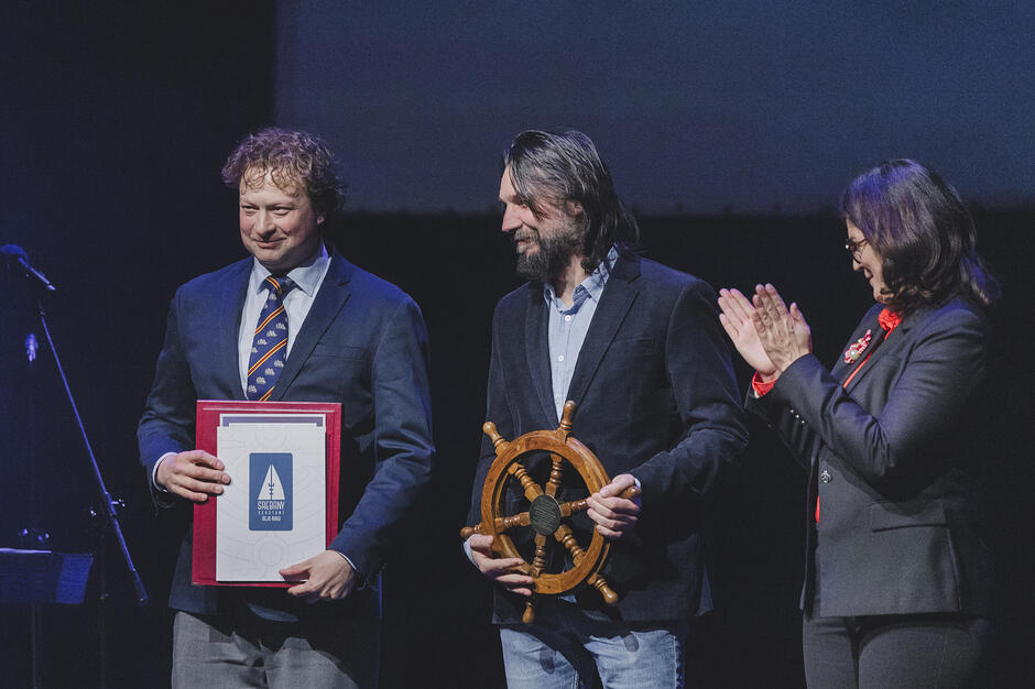 Zdjęcie przedstawia troje uśmiechniętych ludzi na scenie podczas uroczystej gali wręczenia nagród o tematyce morskiej. Mężczyzna po lewej trzyma oprawiony dyplom, centralna postać prezentuje ozdobne koło sterowe, a kobieta po prawej bije im brawo.