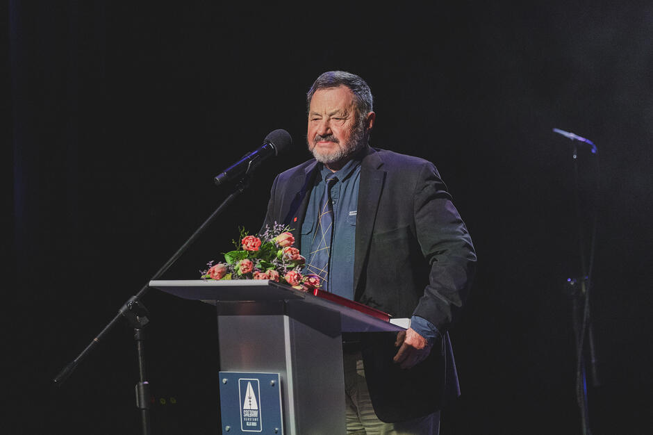 Zdjęcie przedstawia starszego mężczyznę, który przemawia na scenie podczas oficjalnego wydarzenia. Stoi on przy mównicy ozdobionej kwiatami i logo, ubranym w ciemną marynarkę oraz koszulę z krawatem w geometryczne wzory