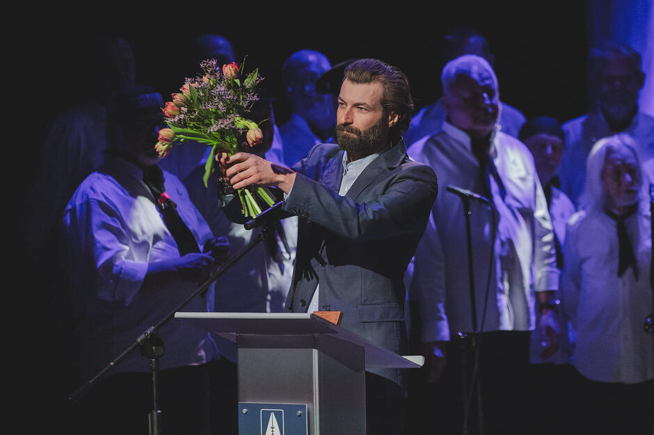 Zdjęcie przedstawia mężczyznę z brodą, w ciemnej marynarce, który trzyma bukiet kwiatów podczas oficjalnego wydarzenia. Znajduje się on przy mównicy z logotypem, a w tle widać grupę osób, co sugeruje uroczystą galę lub ceremonię wręczenia nagród