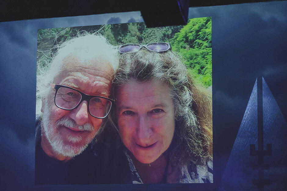 Zdjęcie wyświetlane na ekranie przedstawia uśmiechniętą, dojrzałą parę – siwego mężczyznę w okularach oraz kobietę z okularami przeciwsłonecznymi na głowie. Kadr to bliski, prywatny portret wykonany na tle zielonej roślinności, prawdopodobnie podczas wspólnej podróży. 