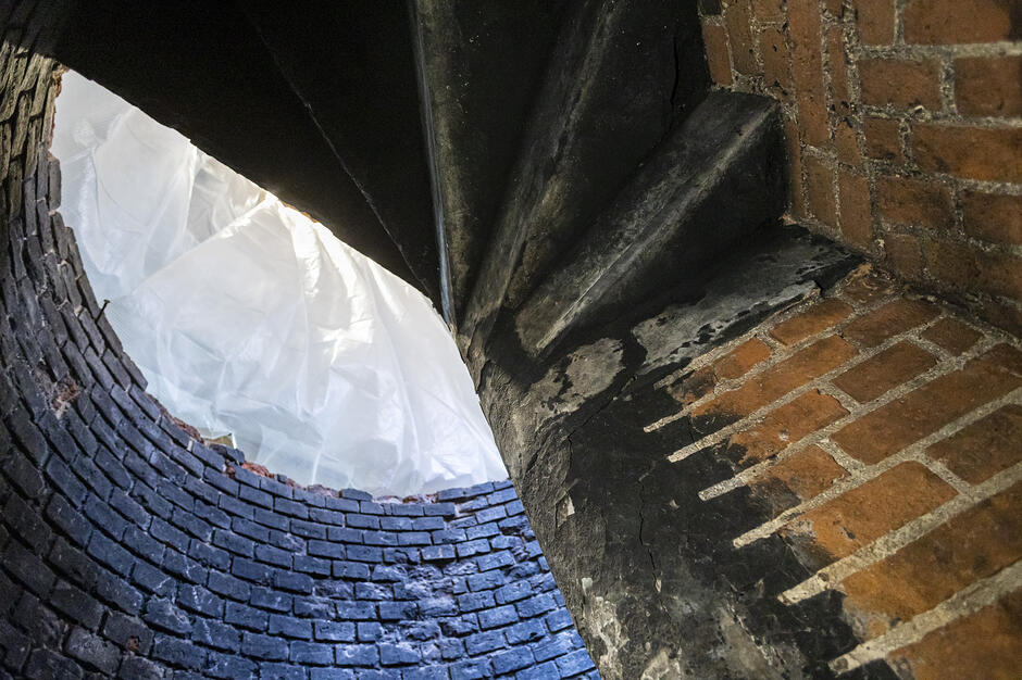 Zdjęcie przedstawia fragment wnętrza starej, ceglanej budowli z krętymi, kamiennymi schodami prowadzącymi w górę. W centralnej części widoczna jest jasna, półprzezroczysta płachta materiału lub folii, która przesłania otwór i wpuszcza do środka rozproszone światło.