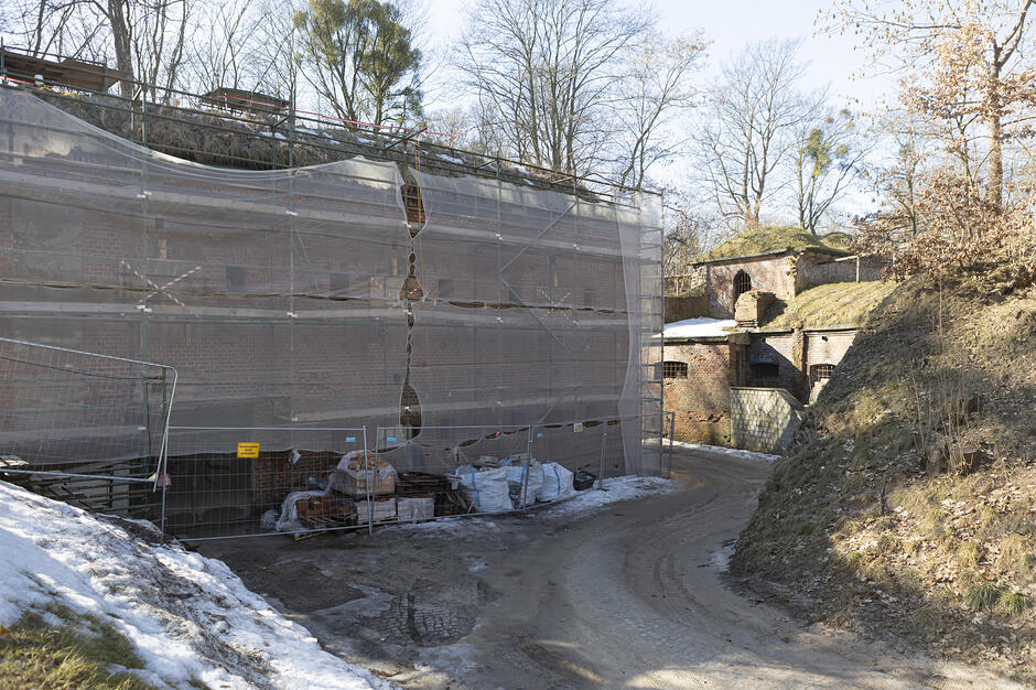 Zdjęcie przedstawia ceglany, historyczny obiekt obronny, którego główna elewacja jest zasłonięta siatką ochronną ze względu na prowadzone prace remontowe. Obok budynku przebiega gruntowa droga, a w otoczeniu widoczne są pagórkowaty teren, resztki śniegu oraz bezlistne drzewa.