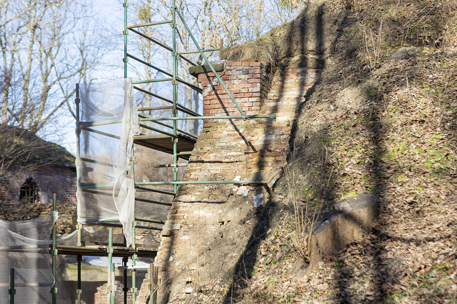 Zdjęcie przedstawia fragment starego, ceglanego muru wkomponowanego w ziemny nasyp, przy którym ustawiono metalowe rusztowania z siatką zabezpieczającą. Widoczne prace renowacyjne odbywają się w otoczeniu drzew i suchej roślinności, sugerując odnawianie historycznego obiektu o charakterze obronnym.