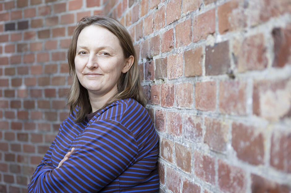 Zdjęcie przedstawia kobietę o jasnych włosach, ubraną w niebiesko-brązową bluzkę w paski, która stoi z założonymi rękami. Postać opiera się o surową ścianę wykonaną z czerwonej cegły, a cała kompozycja utrzymana jest w spokojnym, portretowym stylu. 
