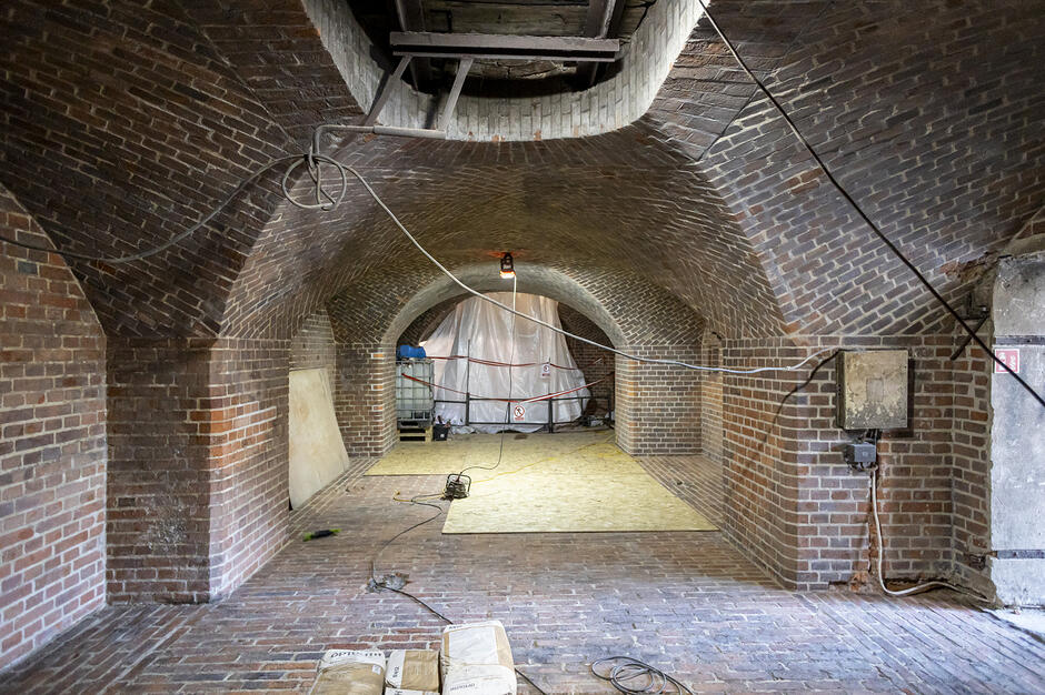 Remont Kaponiery ŚrodkowejZdjęcie przedstawia surowe, ceglane wnętrze z niskimi łukami i sklepieniami, które sprawia wrażenie piwnicy lub dawnego bunkra. W pomieszczeniu trwają prace remontowe, o czym świadczą prowizoryczne oświetlenie, ułożone na podłodze płyty OSB oraz materiały budowlane.