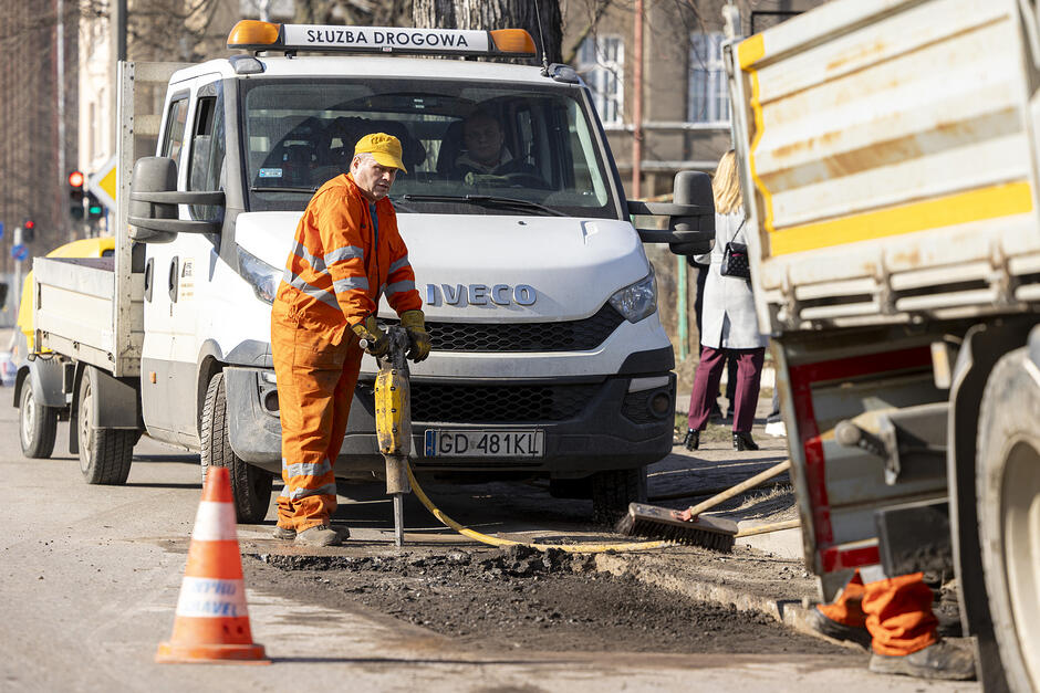 na zdjęciu robotnik w pomarańczowym kombinezonie, wiertarką rozkuwa asfalt w jezdni, za nim stoi samochód dostawczy