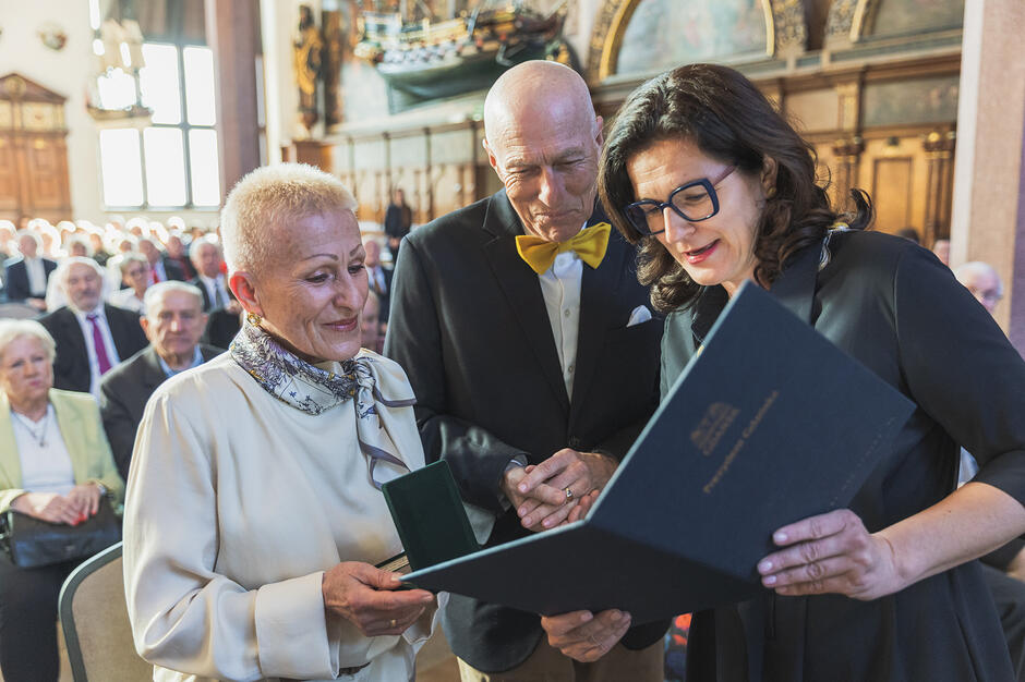 na zdjęciu jedna kobieta trzyma oprawiony dokument w ciemnej okładce, czyta z niego, patrzy na to też starsze małżeństwo